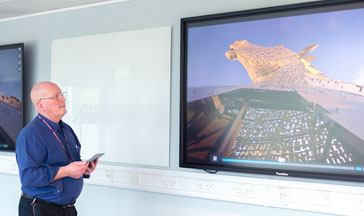 A 33Ƶ staff member standing by 2 screens showing the Scottish Kelpies sculpture