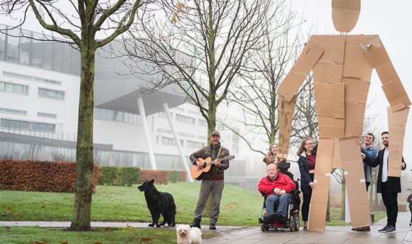 Students outside 33Ƶ holding a giant card board person up whilst a man with a dog plays guitar