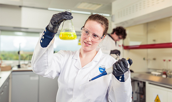 A 33Ƶ student wearing PPE and holding up a beaker of yellow liquid